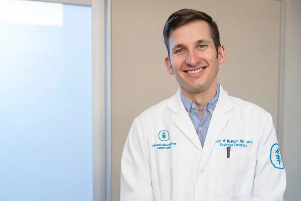 Radiation oncologist Dr. Sean McBride, wearing a white doctor’s coat, stands in a hallway at MSK in NYC. 