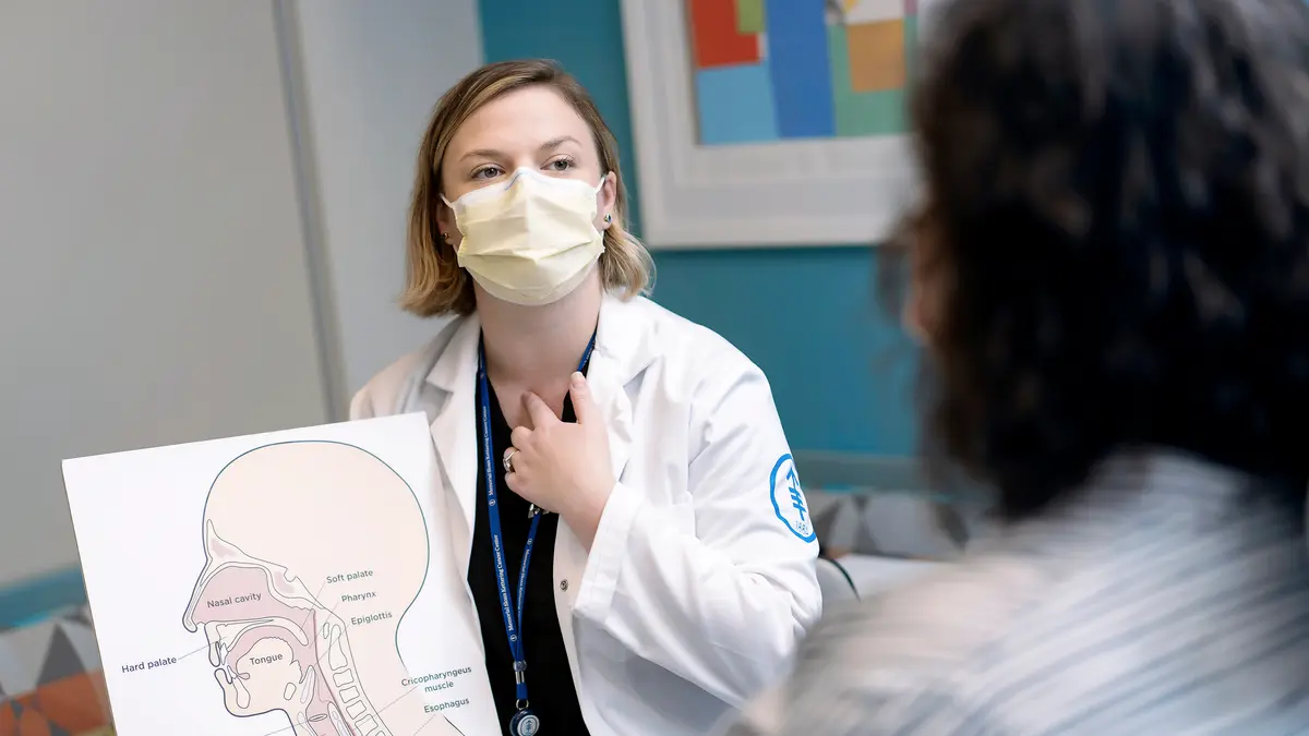 A masked MSK speech pathologist points to her throat as she shows a patient an illustration of the head and neck. 