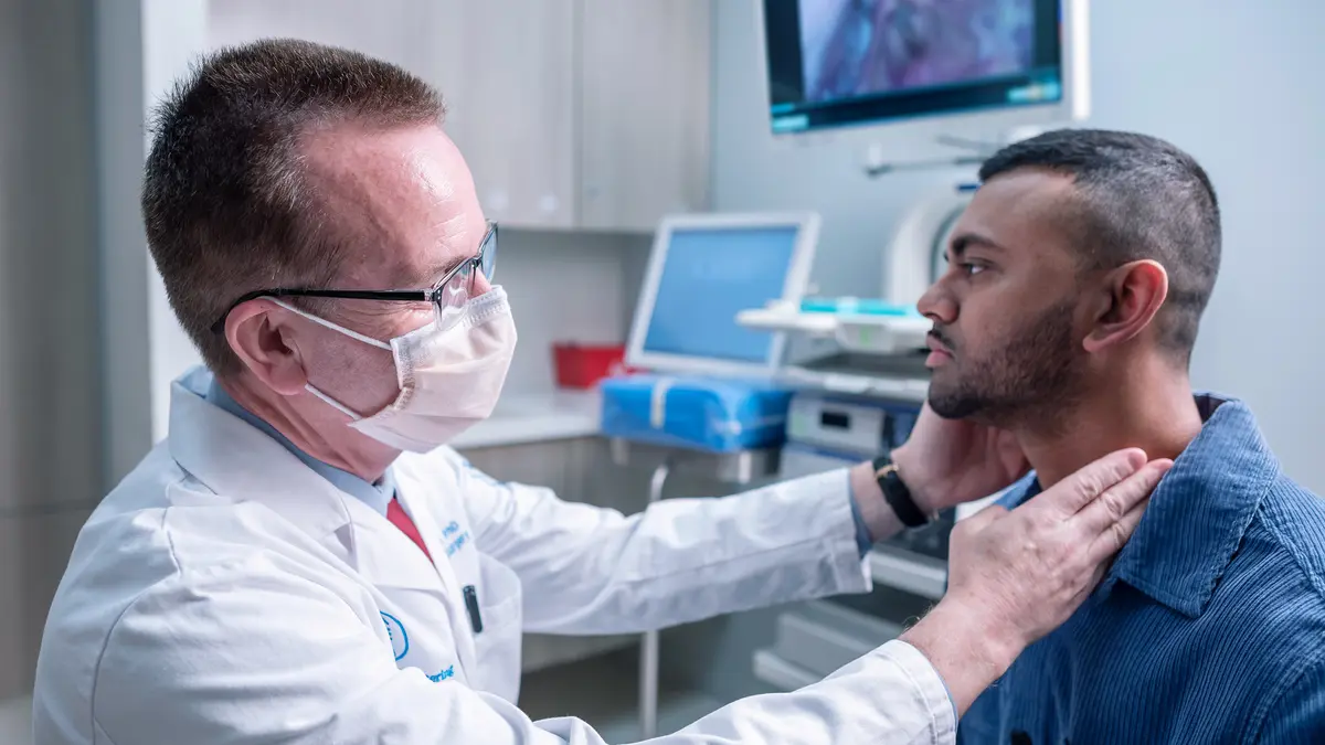Head and neck surgeon Dr. Ian Ganly, wearing a head lamp, feels the throat of his patient in an MSK exam room.  