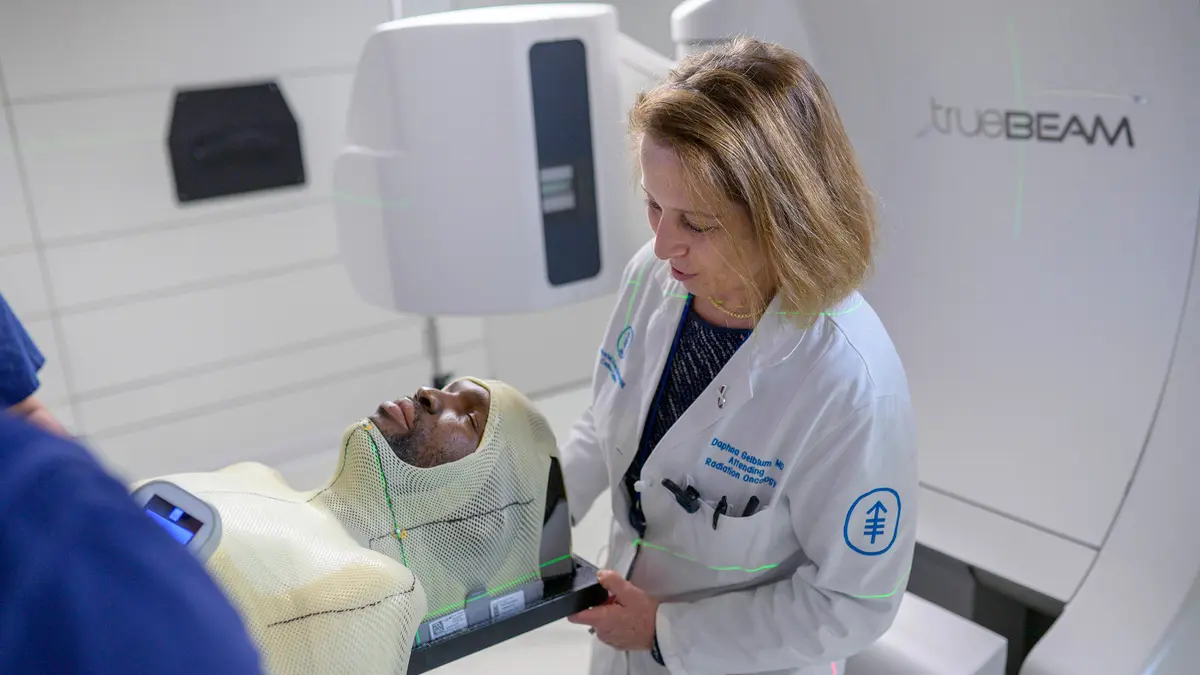 A radiation oncologist is with her patient as he wears a protective mask and lays on a radiation therapy machine. 
