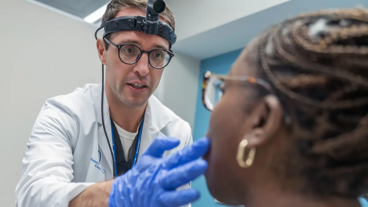 A speech pathologist wearing a headlamp uses his gloved hands to touch a check of his patient. 