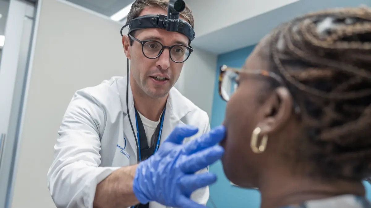 A speech pathologist wearing a headlamp uses his gloved hands to touch a cheek of his patient. 