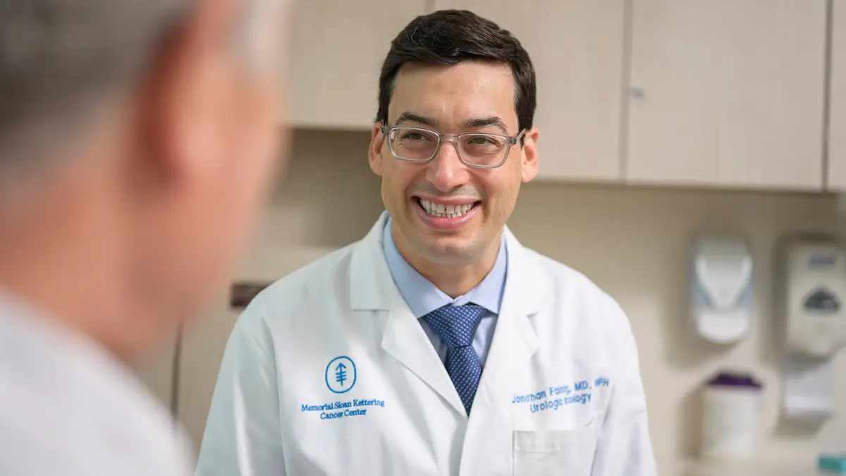 Dr. Fainberg, in a white doctors’ coat, is in an exam room talking with his patient. 