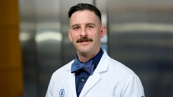 Nicholas Romano,  in a white coat and bow tie, is standing in a hallway at MSK.